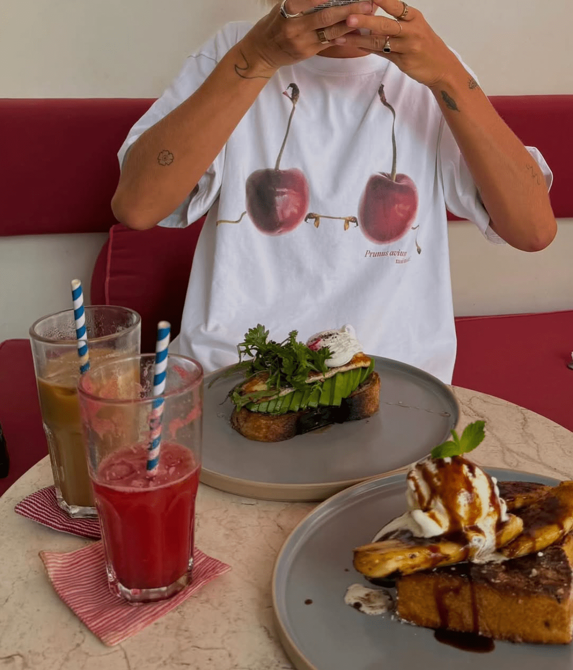 A person at a café table with avocado toast, French toast with ice cream, and two drinks, wearing a cherry-print shirt.